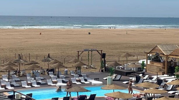 A beachside pool area with lounge chairs and umbrellas, a sign reading "AMIRA BEACH", and a view of the ocean.