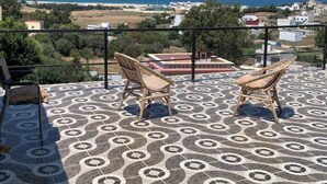 A balcony with patterned flooring, two wicker chairs, and a view of the sea and distant buildings.