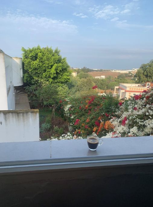 A balcony with a cup of coffee, a view of a garden with flowers, and a building in the background.