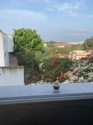 Un balcon avec une tasse de café, une vue sur un jardin fleuri et un bâtiment en arrière-plan.