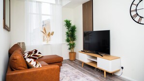 A modern living room with a brown leather sofa, a flat-screen TV on a wooden stand, a large potted plant, and a decorative wall clock.