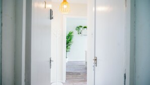 A hallway with white walls, a wooden floor, and a hanging light fixture. There are doors on either side and a plant in the center.