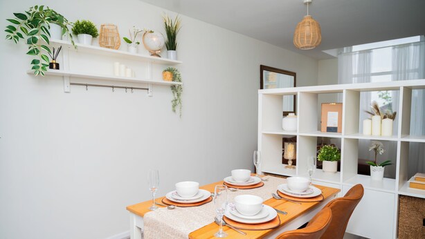 A dining area with a long table set for four, white shelves with plants, and a hanging light fixture.