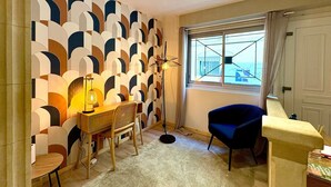 Ein Zimmer mit einer Wand mit geometrischem Muster, einem Schreibtisch mit Stuhl, einem blauen Sessel, einem Fenster mit Meerblick und einer Tür.