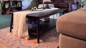 Un salón con un sofá de cuero marrón oscuro, un sillón blanco, una mesa de centro con libros, un televisor montado en la pared y una escalera al fondo.