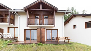 A two-story wooden house with a balcony, large windows, and a wooden deck with outdoor furniture.