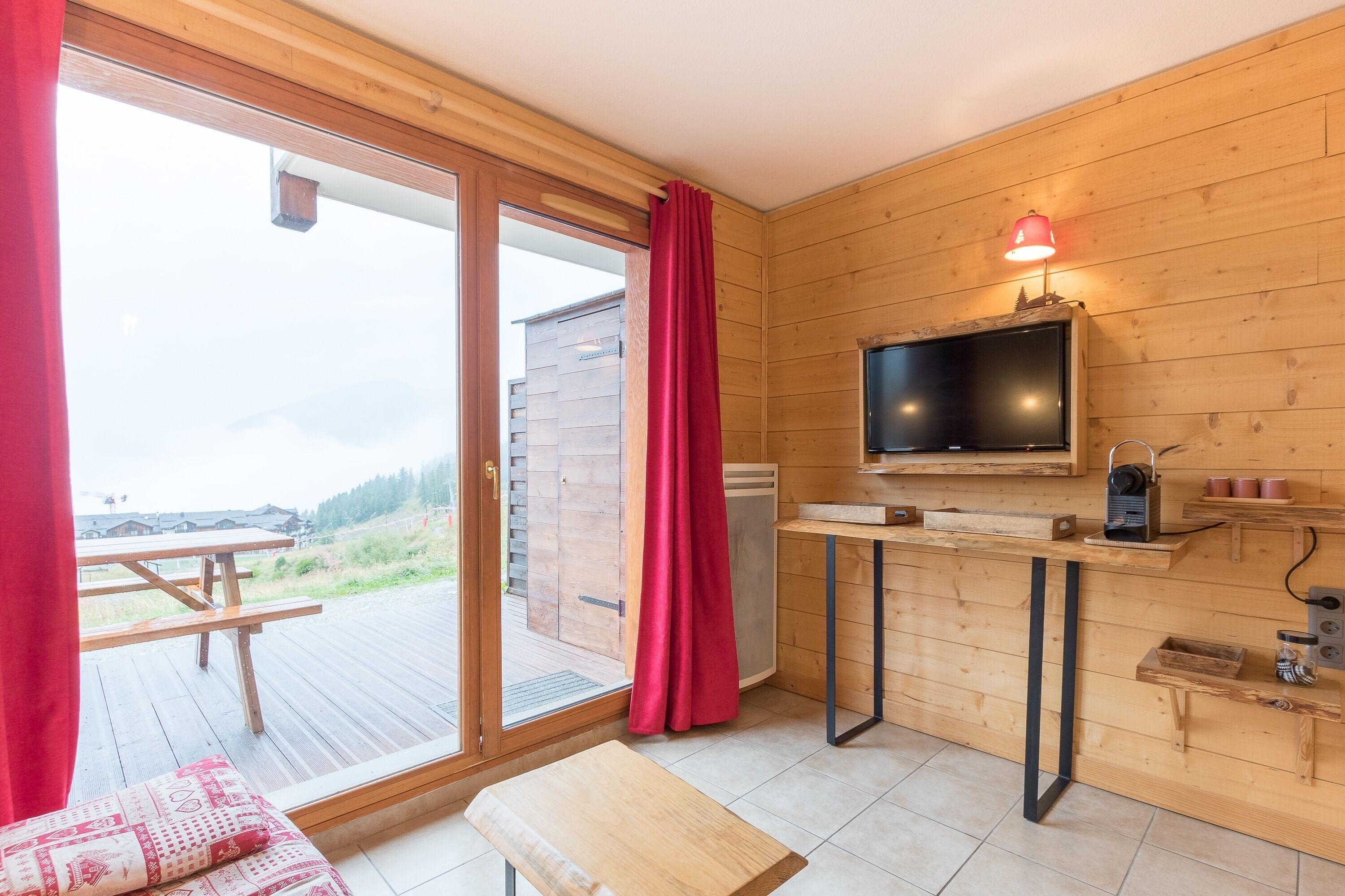A wooden interior with a sliding glass door leading to an outdoor deck, a small kitchenette with a TV, and a bench.