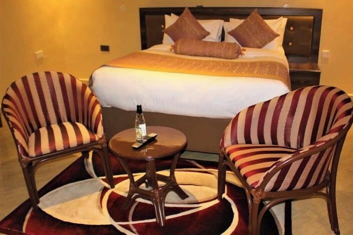 A hotel room with a large bed, two striped armchairs, a small round table, and a red carpet.