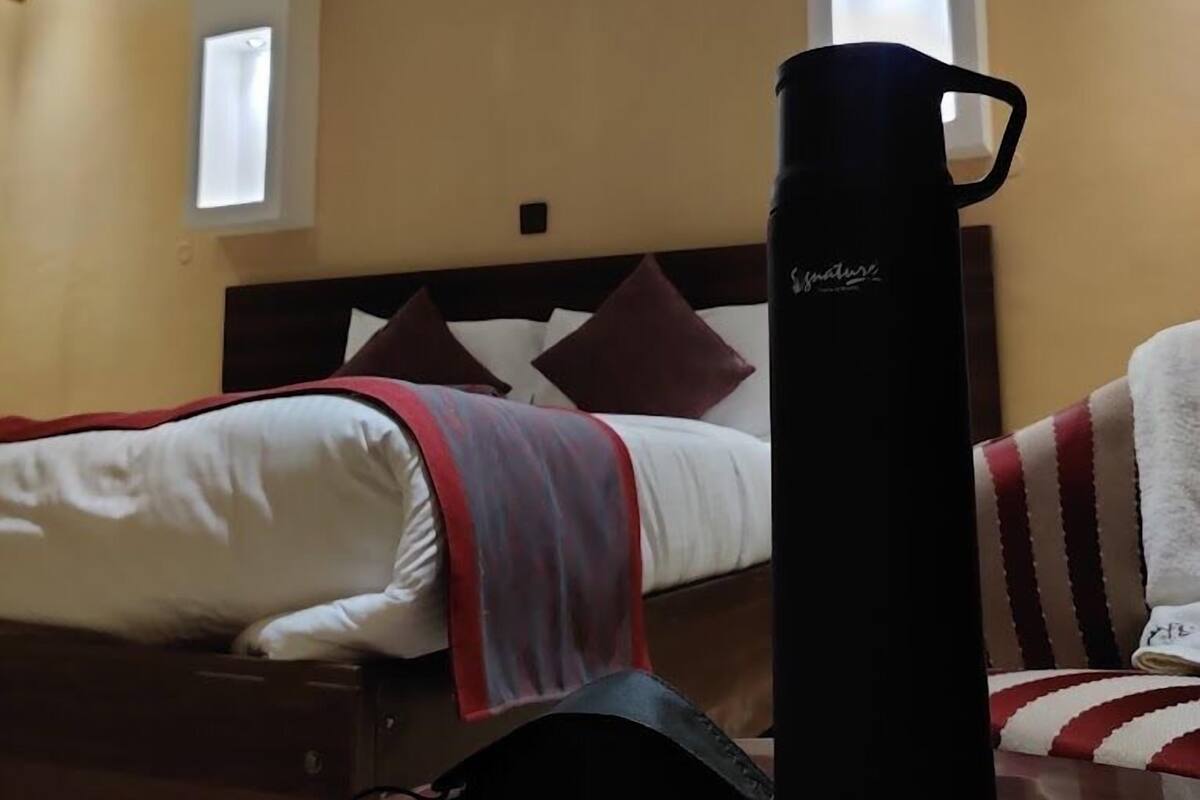 A hotel room with a bed, a bedside table, a black water bottle, and a window.