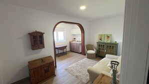 Una habitación con un armario de madera, una silla de mimbre, un sofá, una mesita y una cocina visible a través de un arco.