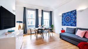 A modern living room with a grey sofa, a dining table with chairs, a TV on a white cabinet, and a large blue artwork on the wall.