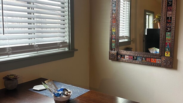 Een kamer met een raam met jaloezieën, een spiegel met decoratieve tegels, een tafel met tafelonderzetter, en een stopcontact aan de muur.