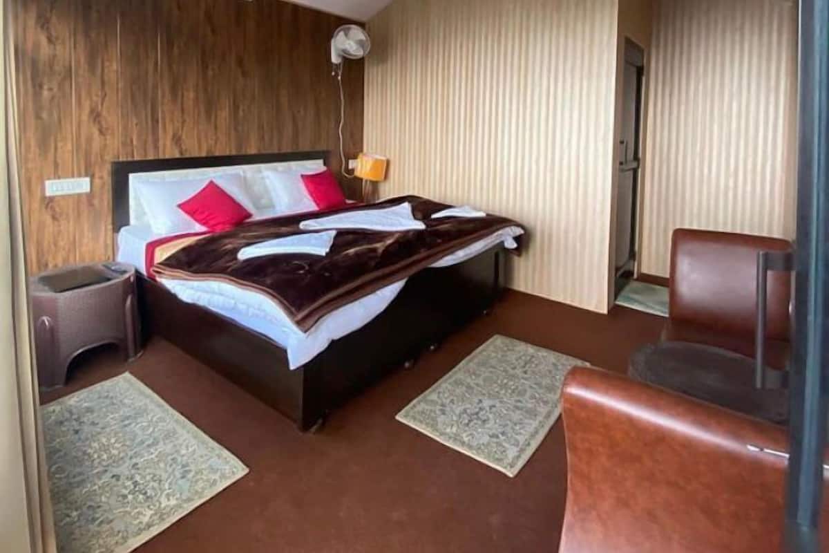 Ein Schlafzimmer mit einem Bett, Nachttischen, einem Sessel, einem Standventilator und einer Deckenleuchte.