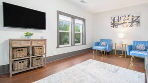 A living room with a flat-screen TV, wicker baskets, blue armchairs, a white rug, and a large abstract painting. - Dunes Getaway! Plush Mattresses! Near Beach! (Michigan City)