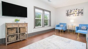 Una sala de estar con una televisión de pantalla plana, cestas de mimbre, sillones azules, un tapete blanco y un ventanal grande.