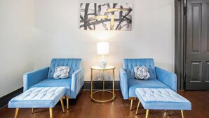 Una habitación con dos sillones azules, una mesita redonda con una lámpara y un cuadro abstracto grande en la pared.
