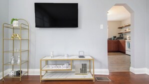 Ein Wohnzimmer mit an der Wand montiertem Fernseher, einem goldfarbenen Konsolentisch mit Marmorplatte und einer kleinen Ablage mit dekorativen Gegenständen.