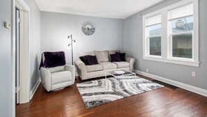 Ein Wohnzimmer mit einer Couch, einem Sessel und einem Couchtisch, einer Uhr an der Wand und einem Fenster mit Jalousien.
