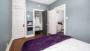 Ein Schlafzimmer mit einem Bett, einer violetten Tagesdecke, einem Kleiderschrank mit Kleidung und einer Tür zu einem anderen Raum.