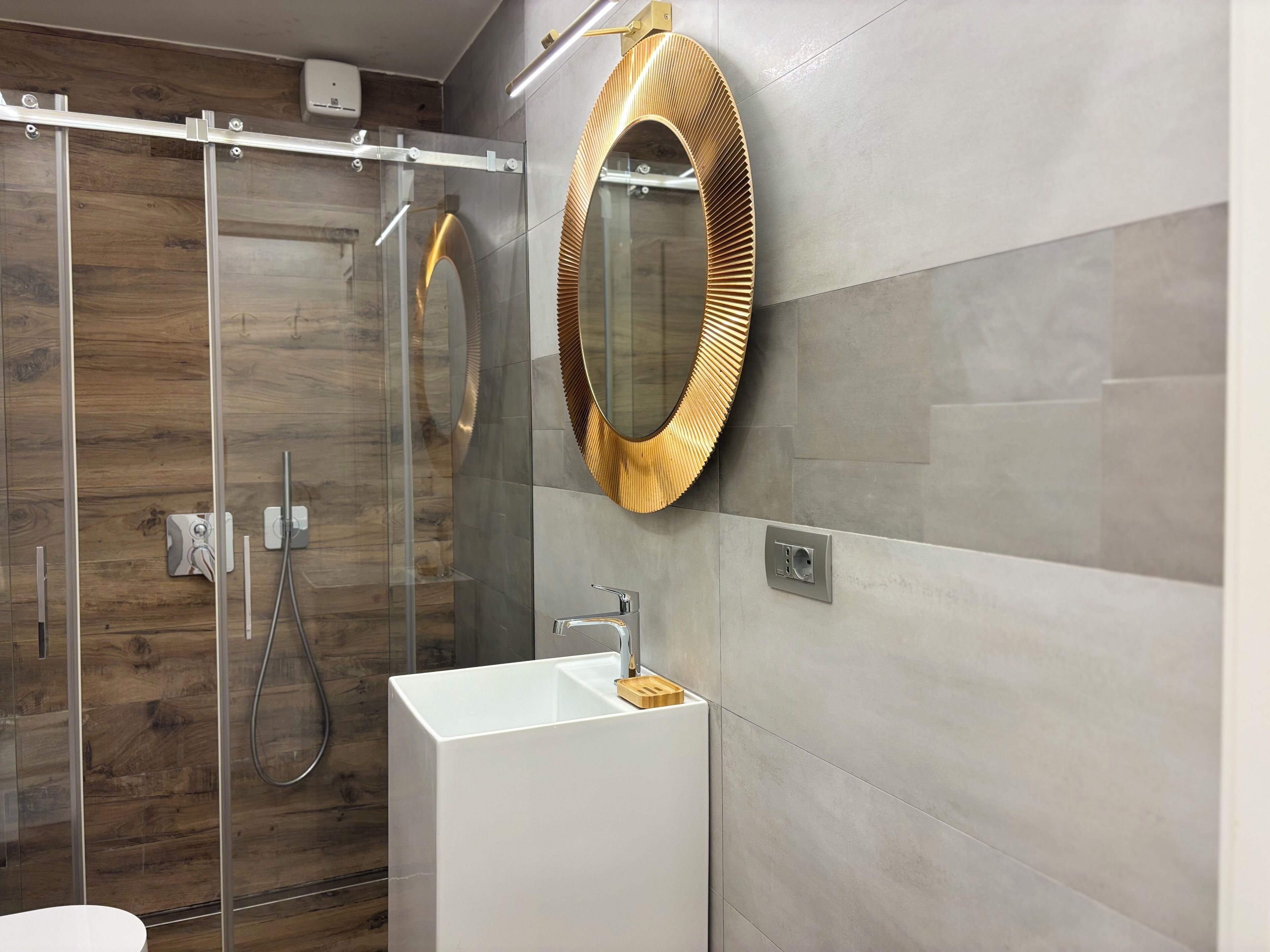 Un baño moderno con cabina de ducha de vidrio, un lavabo blanco y un espejo con detalles dorados.