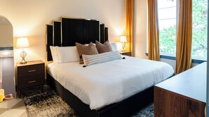Un dormitorio moderno con una cama grande, dos mesitas de noche con lámparas, un mueble de noche de madera, un cabecero negro y vistas a zonas verdes a través de la ventana.