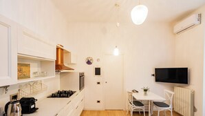 Een kleine keuken met witte kastjes, een fornuis, een koffiezetapparaat en een eethoek met een tafel en stoelen.