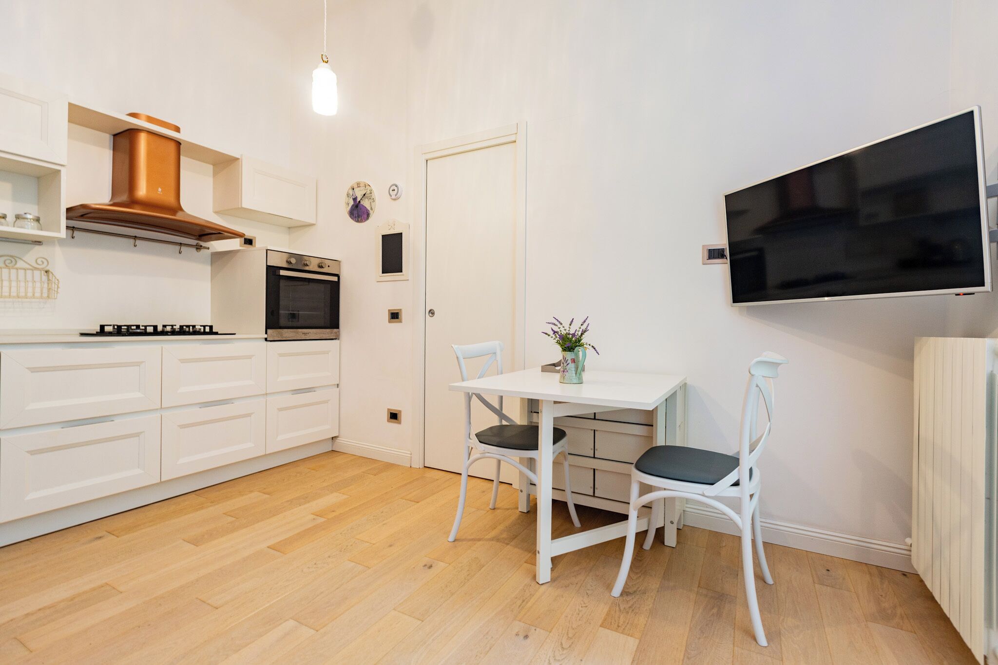 Een moderne keuken met witte kastjes, een koperen afzuigkap, een ingebouwde oven en een eethoek met een tafel en twee stoelen.