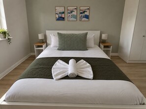 Un lit bien fait, avec une serviette pliée, une couverture verte matelassée et un oreiller blanc. On y trouve deux tables de chevet avec des lampes, une fenêtre avec une plante et trois tableaux abstraits accrochés au mur.