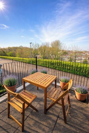Une table et des chaises en bois sur un balcon, agrémenté de pots de lavande, avec une clôture en métal et une vue sur un quartier résidentiel.