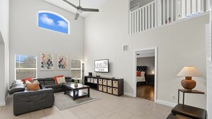 A spacious living room with a black leather sofa, a coffee table, and a TV mounted on the wall. There is a ceiling fan, a window with blinds, and a staircase leading to an upper level.