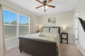 Ein Schlafzimmer mit einem Bett, einem Deckenventilator, einem Fenster mit Jalousien, einem Nachttisch und einem Bild an der Wand.