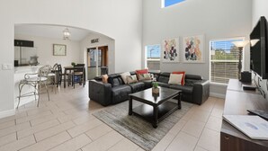 A spacious living room with a black leather sofa, a coffee table, and a dining area with a table and chairs. There is a kitchen area with cabinets and a refrigerator. The room has large windows with blinds and a ceiling with a skylight.