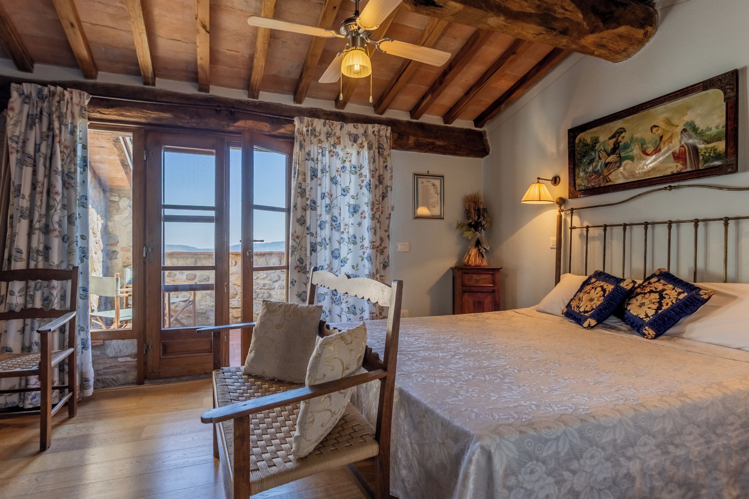 A bedroom with a bed, a chair, a table, a ceiling fan, and a painting on the wall.