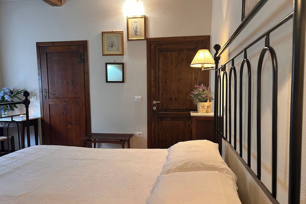 A bedroom with a bed, a desk, and a chair. There are two doors, one with a wooden frame and one with a metal frame. A lamp is on the desk.