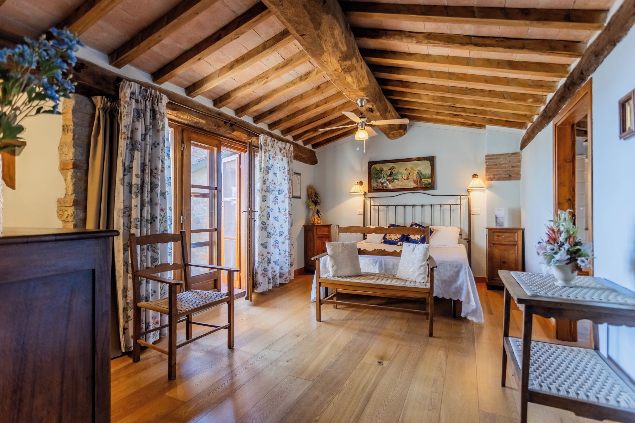 Una habitación con techo de madera, una cama con cabecera, una silla, una mesa y un ventanal con cortinas.