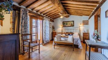 Una habitación con techo de madera, una cama con cabecera, una silla, una mesa y un ventanal con cortinas.