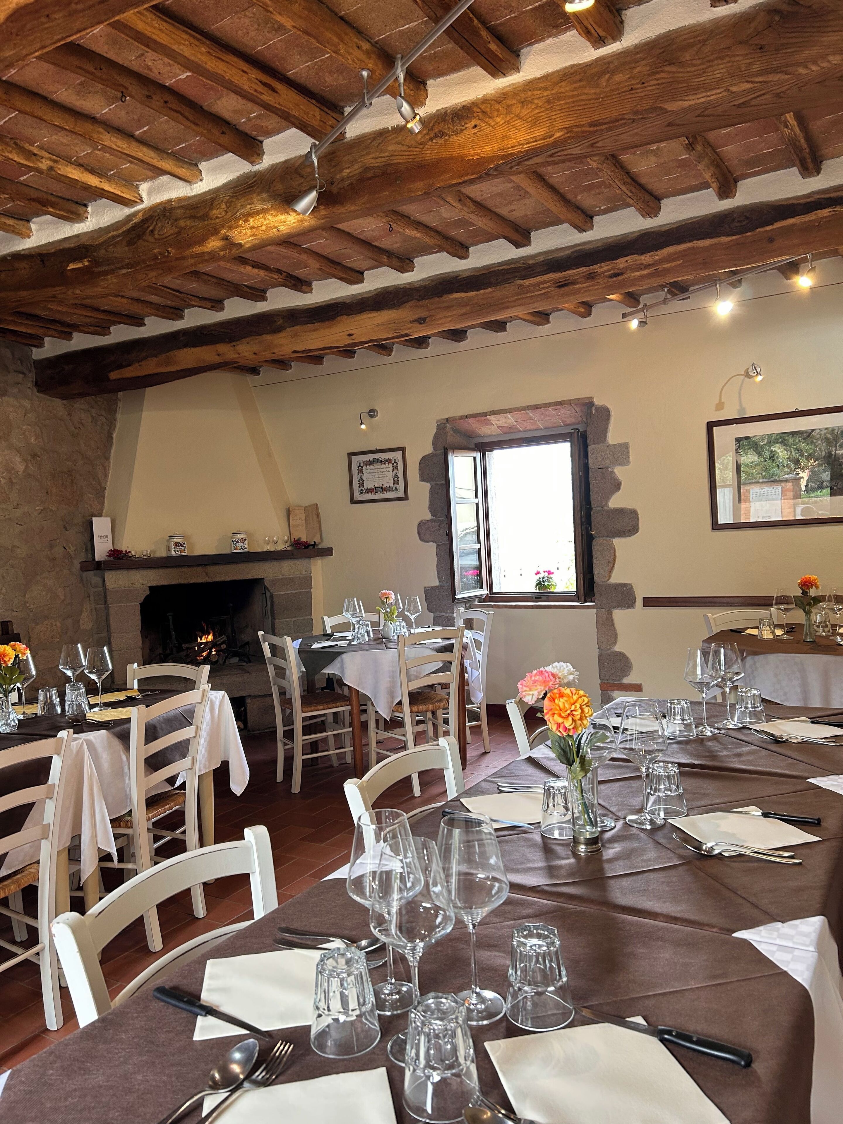 A dining area with wooden beams, stone walls, and a fireplace. Tables are set with wine glasses and cutlery.