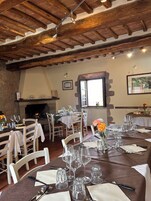 A dining area with wooden beams, stone walls, and a fireplace. Tables are set with wine glasses and cutlery.