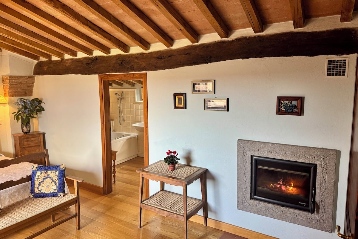 A cozy living room with a fireplace, wooden beams, a sofa, a small table, and a view into the bathroom.