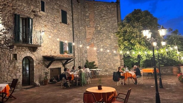 Un espacio al aire libre para comer con edificios de piedra, mesas con manteles anaranjados y luces colgantes.