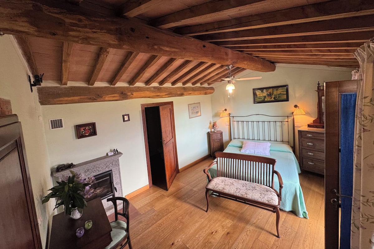 A rustic bedroom with exposed wooden beams, a bed with a floral bedspread, a wooden chair, a fireplace, and a dresser.