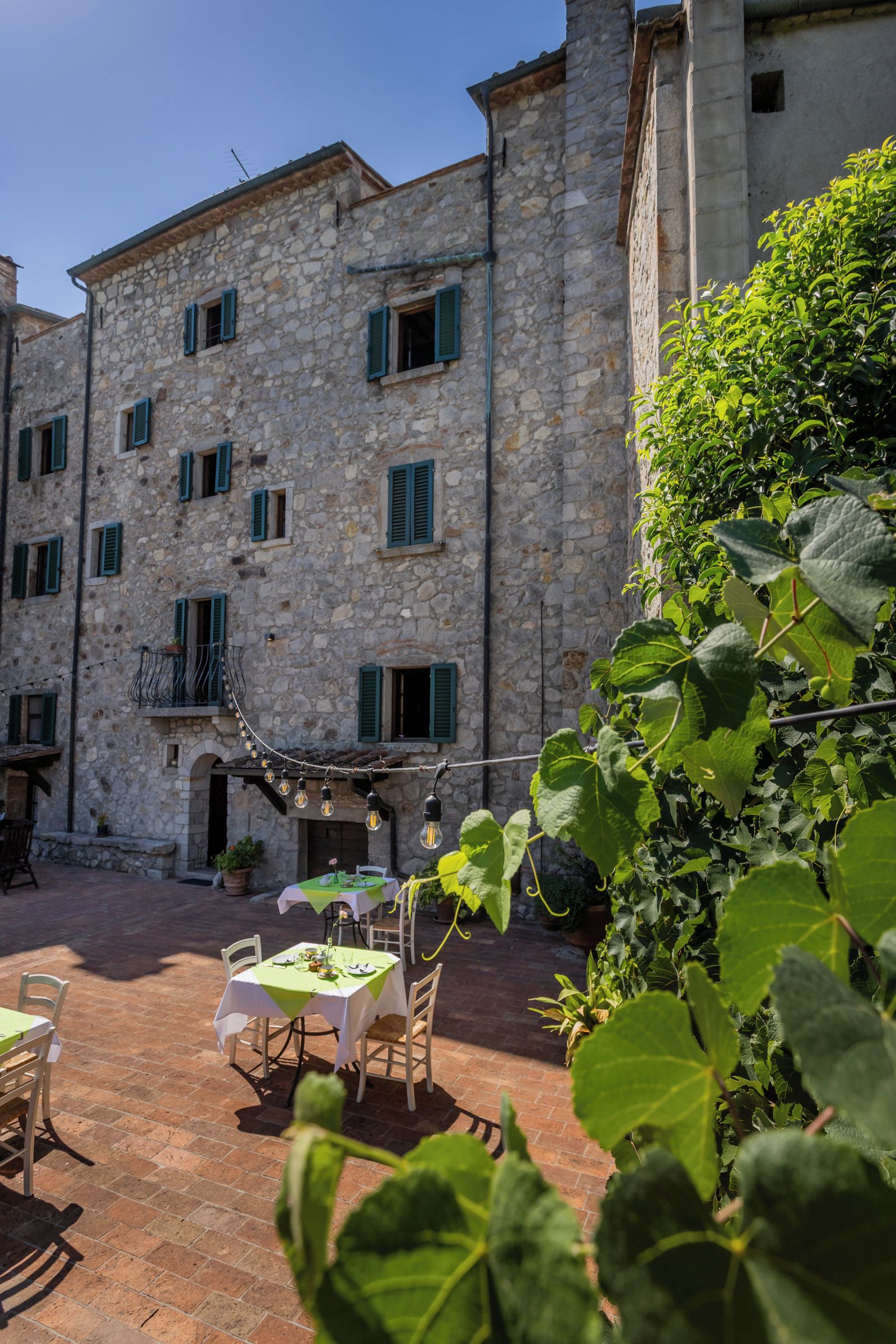 A stone building with multiple windows and a balcony, a table set for dining, and lush greenery in the foreground.