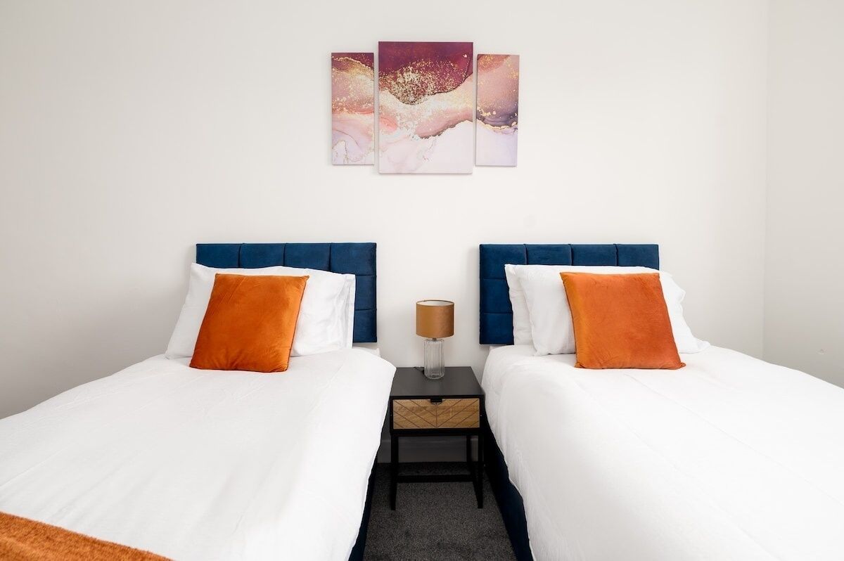 Et hotelværelse med to senge, hvidt sengetøj, blå sengegavl og orange puder. Der er et natbord med en lampe og en lille skuffe.