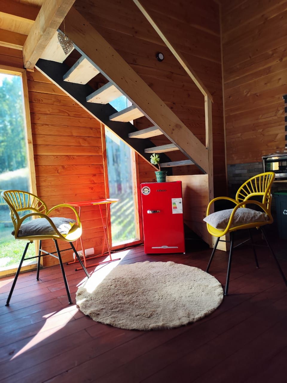 Interior de una cabaña de madera con techo alto, un refrigerador rojo, dos sillas amarillas, una alfombra blanca y una ventana con vistas a la vegetación.