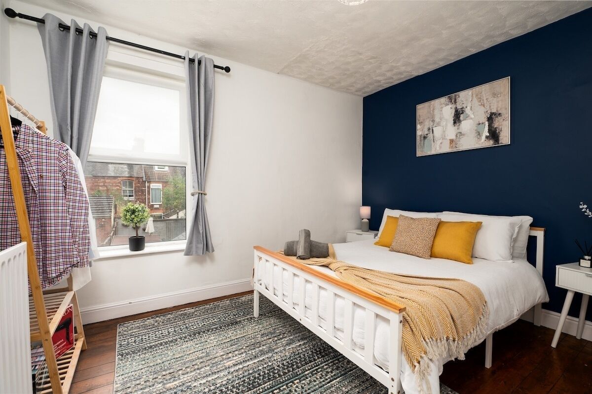 A bedroom with a bed, a window with curtains, a radiator, a bedside table, and a painting on the wall.