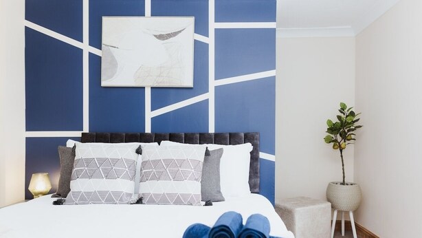 A neatly made bed with geometric patterned pillows, a blue towel, and a framed abstract artwork on the wall. - Stunning House in Heart of Historic Lincoln (Lincolnshire)