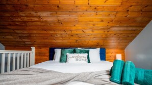Ein Schlafzimmer mit Holzboden, einem Bett mit weißer Bettwäsche, türkisfarbenen Kissen und einem Kissen mit der Aufschrift 'Willkommen Zuhause'.