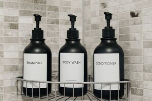 Tres botellas negras de champú, gel de baño y acondicionador sobre una estantería metálica frente a una pared de azulejos.