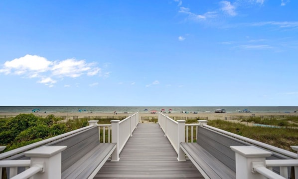 A wooden boardwalk leading to a beach with umbrellas and people in the distance.