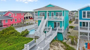 A multi-story building with a prominent staircase leading to a balcony, surrounded by other colorful houses. - The Perfect Oceanfront Vacation w/ Private Pool! (Holden Beach)